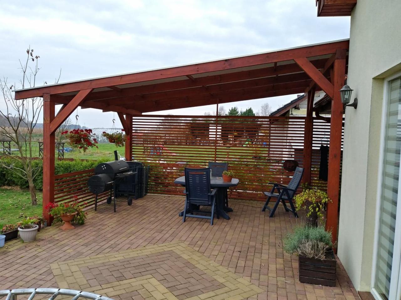 Zadaszony taras z drewnianą konstrukcją, wyposażony w stół, krzesła i grill, z dekoracyjnymi donicami z kwiatami na ceglanej posadzce.