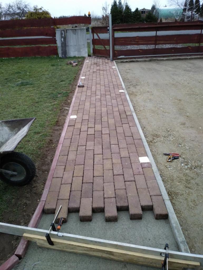 Układanie czerwonej kostki brukowej na podjeździe, widoczne narzędzia, poziomica i taczka, etap prac przed ostatecznym wyrównaniem.