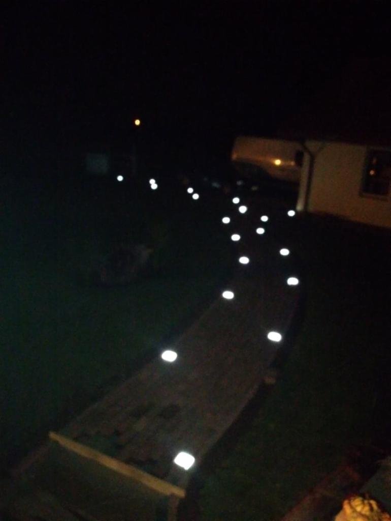 Oświetlona ścieżka w ogrodzie nocą, z lampami punktowymi wzdłuż drewnianego chodnika prowadzącego do domu.