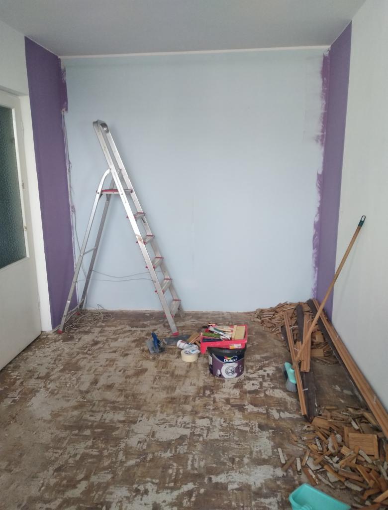 Pokój w trakcie przygotowania do malowania: ściany częściowo pomalowane na fioletowo, drabina aluminiowa, wiadro z farbą Dulux, resztki starej podłogi na podłodze.