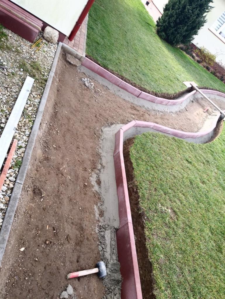 Układanie obrzeży trawnikowych z czerwonej kostki, widoczny młotek i zaprawa cementowa, trawa po prawej stronie.