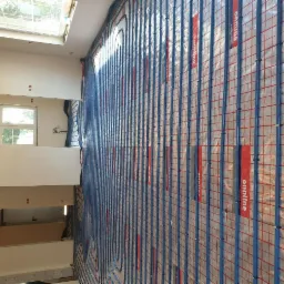 Niebieskie rurki ogrzewania podłogowego ułożone na siatce montażowej w jasnym pomieszczeniu z oknem dachowym i widoczną instalacją elektryczną w ścianie.