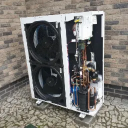 Rozłożona, biała obudowa pompy ciepła z widocznymi dwoma wentylatorami, miedzianymi rurami i elementami elektronicznymi na tle ceglanej ściany i kamiennej kostki brukowej.