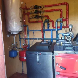 Kompleksowa instalacja grzewcza z piecem na paliwo stałe, zbiornikiem buforowym i licznymi rurami w izolacji czerwonej i niebieskiej, widoczne manometry i pompy obiegowe.