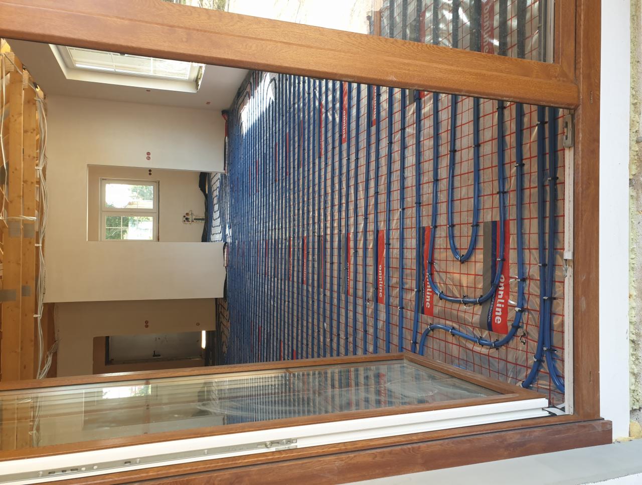 Montaż ogrzewania podłogowego: niebieskie rurki na folii z siatką, widoczne przez drewnianą ramę okienną, w tle niezakończone ściany i okno dachowe.