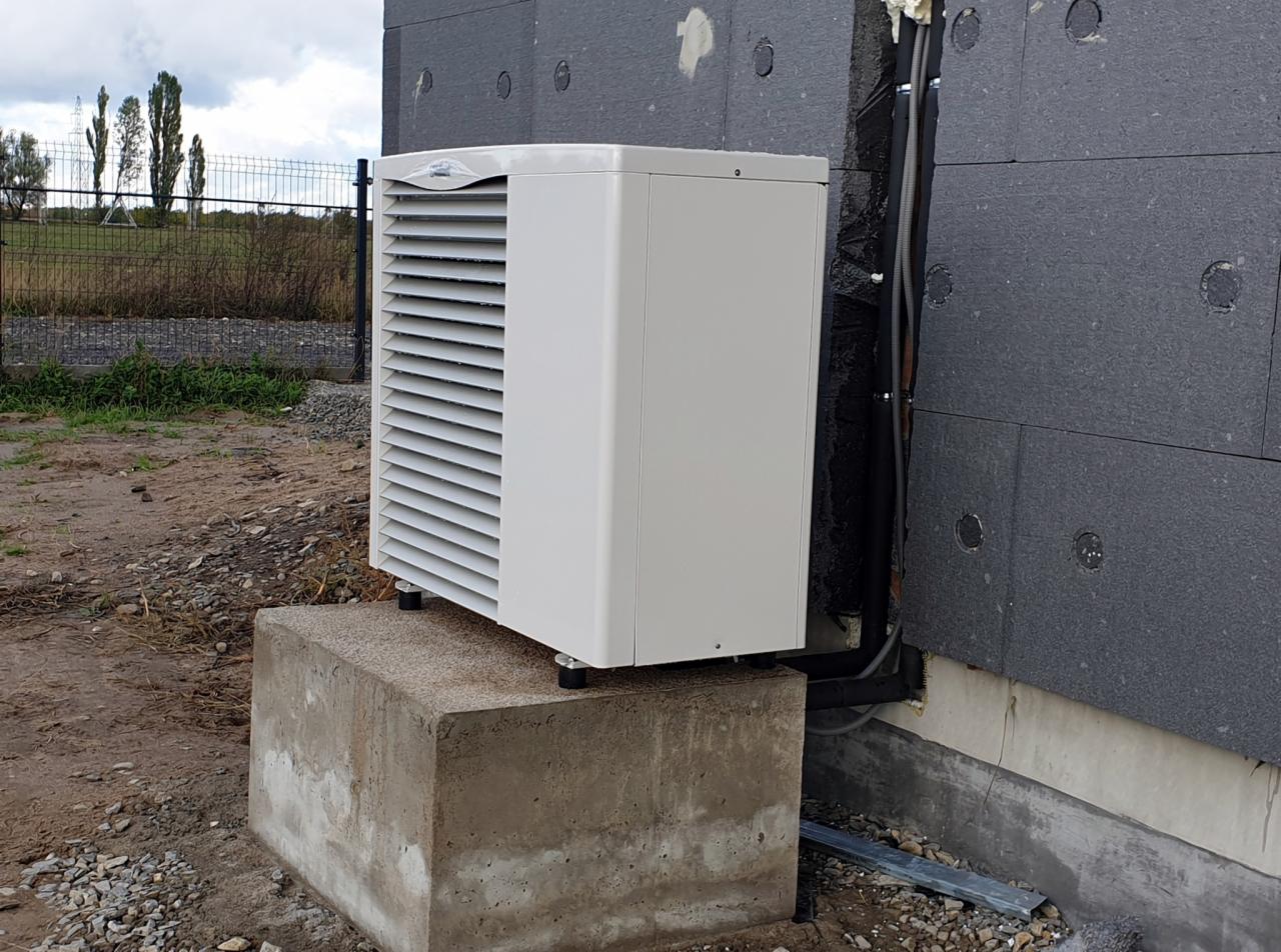 Zewnętrzna jednostka pompy ciepła zamontowana na betonowym fundamencie przy ścianie budynku z szarym ociepleniem, widoczne rury instalacyjne i teren wokół.