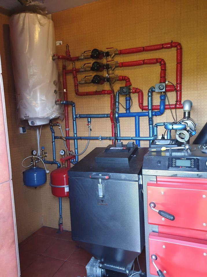 Kompleksowa instalacja grzewcza z piecem na paliwo stałe, zbiornikiem buforowym i licznymi rurami w izolacji czerwonej i niebieskiej, widoczne manometry i pompy obiegowe.