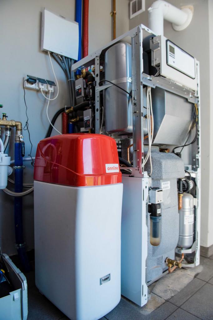 Rozbudowana instalacja grzewcza z zmiękczaczem wody BWT z czerwonym wieczkiem Onnline, widoczne liczne rury, zawory i przewody elektryczne.