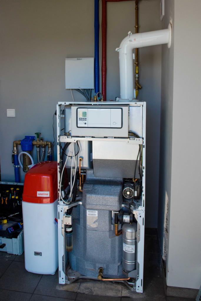 Otwarta obudowa kotła grzewczego z widocznymi elementami wewnętrznymi, obok zmiękczacz wody z czerwonym zbiornikiem i niebieskimi filtrami oraz skrzynka z narzędziami.