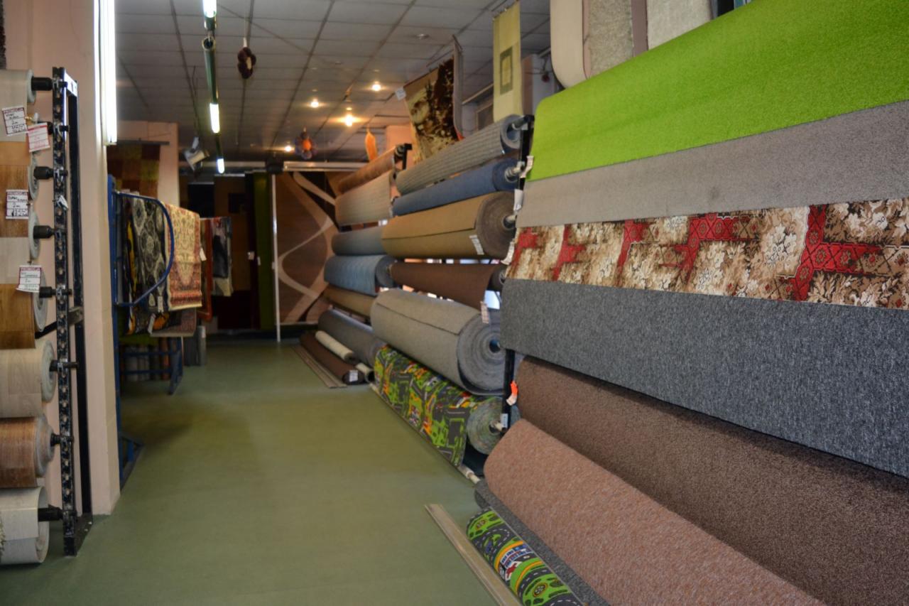 Rzędy zwiniętych dywanów o różnych wzorach i kolorach w magazynie, widoczne rolki z etykietami cenowymi oraz fragmenty innych dywanów w tle.