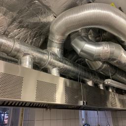 Niko-Tech - Srebrne, izolowane rury wentylacyjne w budynku, przymocowane do metalowego okapu z perforowanymi panelami, widoczne płytki na ścianie i okno w tle.