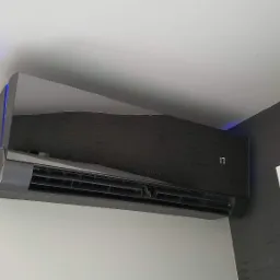 Czarny, nowoczesny klimatyzator z niebieskim podświetleniem LED zamontowany na białej ścianie, widoczny fragment okna z roletą i dekoracyjny element na suficie.