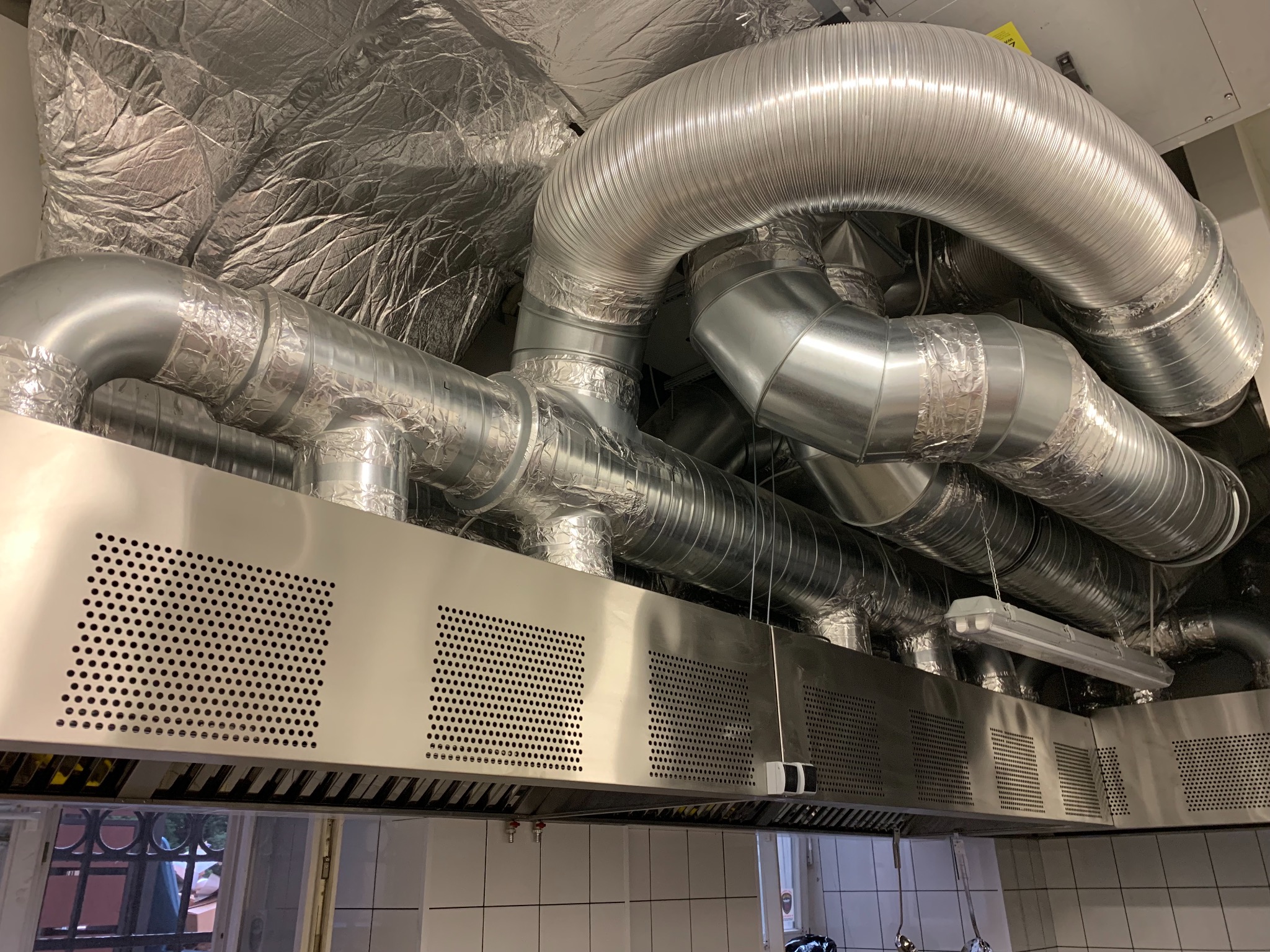 Srebrne, izolowane rury wentylacyjne w budynku, przymocowane do metalowego okapu z perforowanymi panelami, widoczne płytki na ścianie i okno w tle.