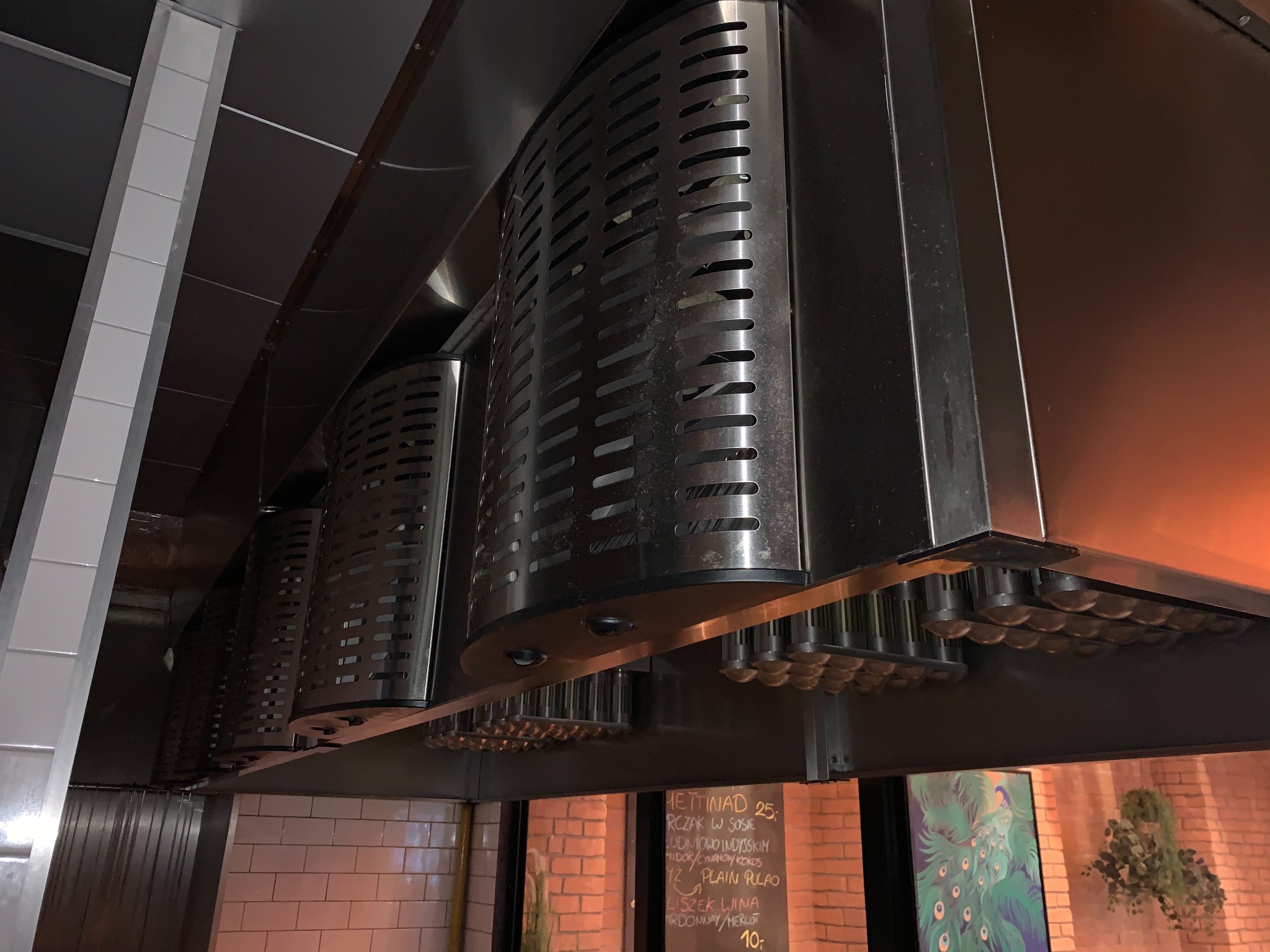 Szereg przemysłowych okapów wentylacyjnych ze stali nierdzewnej, zamontowanych w restauracji, widok od dołu, z widoczną tablicą menu w tle.