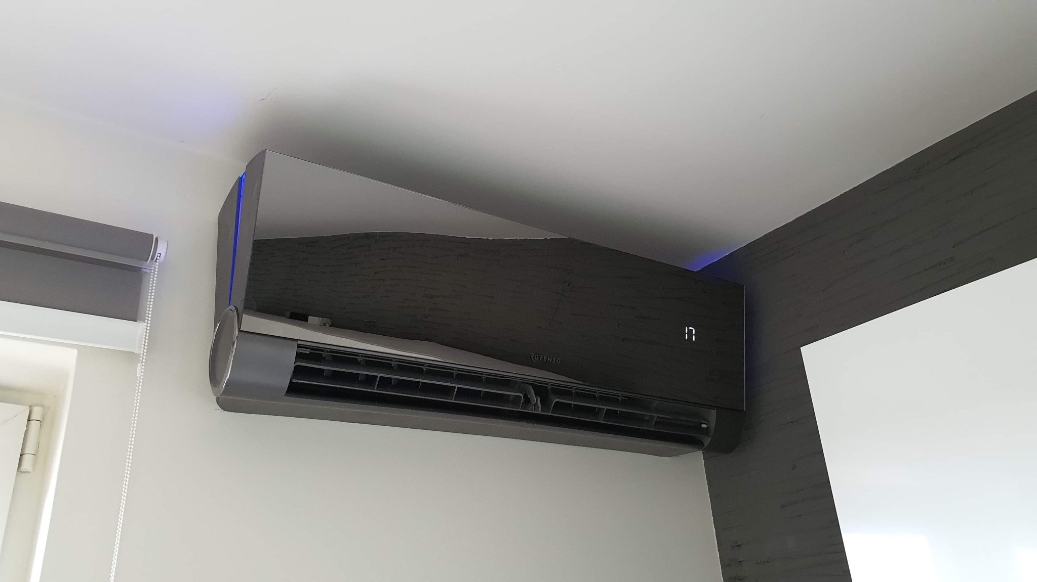 Czarny, nowoczesny klimatyzator z niebieskim podświetleniem LED zamontowany na białej ścianie, widoczny fragment okna z roletą i dekoracyjny element na suficie.