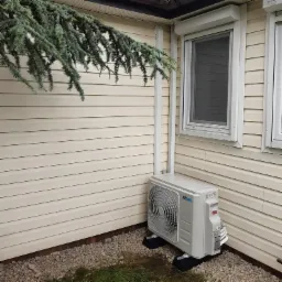 Zewnętrzna jednostka klimatyzacji zamontowana przy ścianie budynku z sidingu, widoczne rury prowadzące do góry, pod jednostką kamienny żwir i fragment trawy, nad jednostką zwisająca gałąź drzewa...