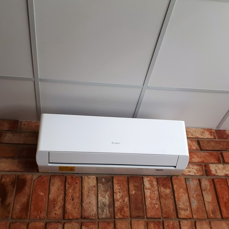 Biała jednostka klimatyzacji marki Gree zamontowana na ceglanej ścianie pod sufitem kasetonowym.