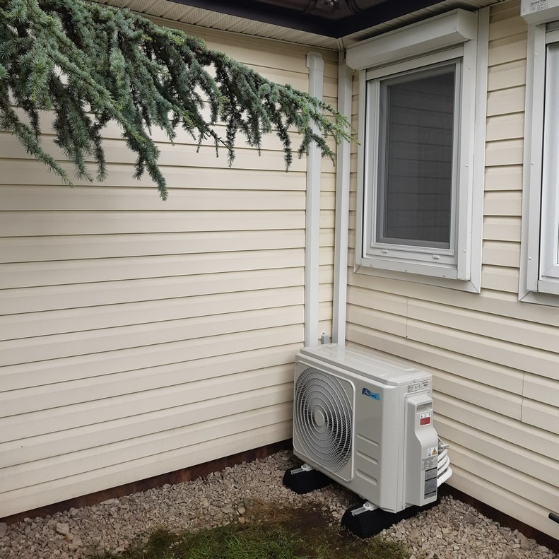 Zewnętrzna jednostka klimatyzacji zamontowana przy ścianie budynku z sidingu, widoczne rury prowadzące do góry, pod jednostką kamienny żwir i fragment trawy, nad jednostką zwisająca gałąź drzewa...