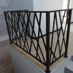 Nowoczesna, kuta balustrada schodowa w kolorze ciemnego brązu, z geometrycznym wzorem krzyżujących się ukośnych prętów, zamontowana na białych schodach w jasnym wnętrzu.