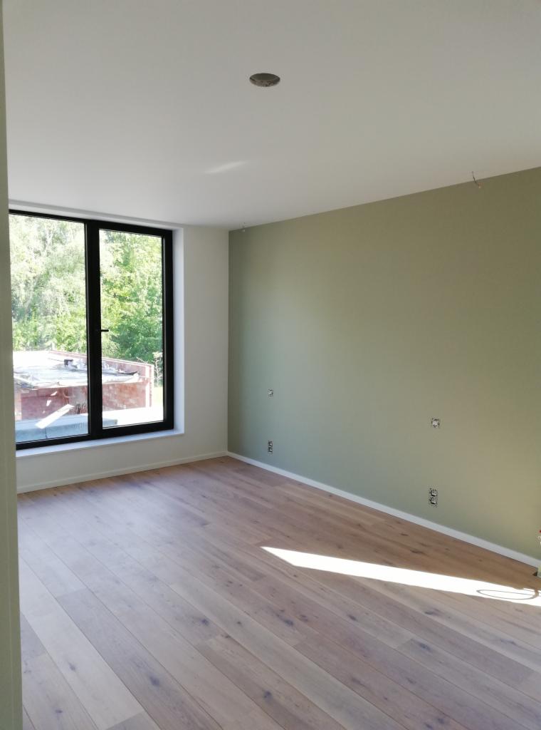 Wnętrze pokoju po remoncie: świeżo pomalowana oliwkowa ściana z widocznymi puszkami elektrycznymi, jasna drewniana podłoga i duże okno z widokiem na zieleń.