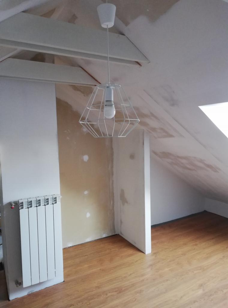 Poddasze w trakcie przygotowania do prac wykończeniowych: ściany z widocznymi śladami po łatach, surowy strop, biały grzejnik i nowoczesna lampa wisząca z metalowym kloszem.