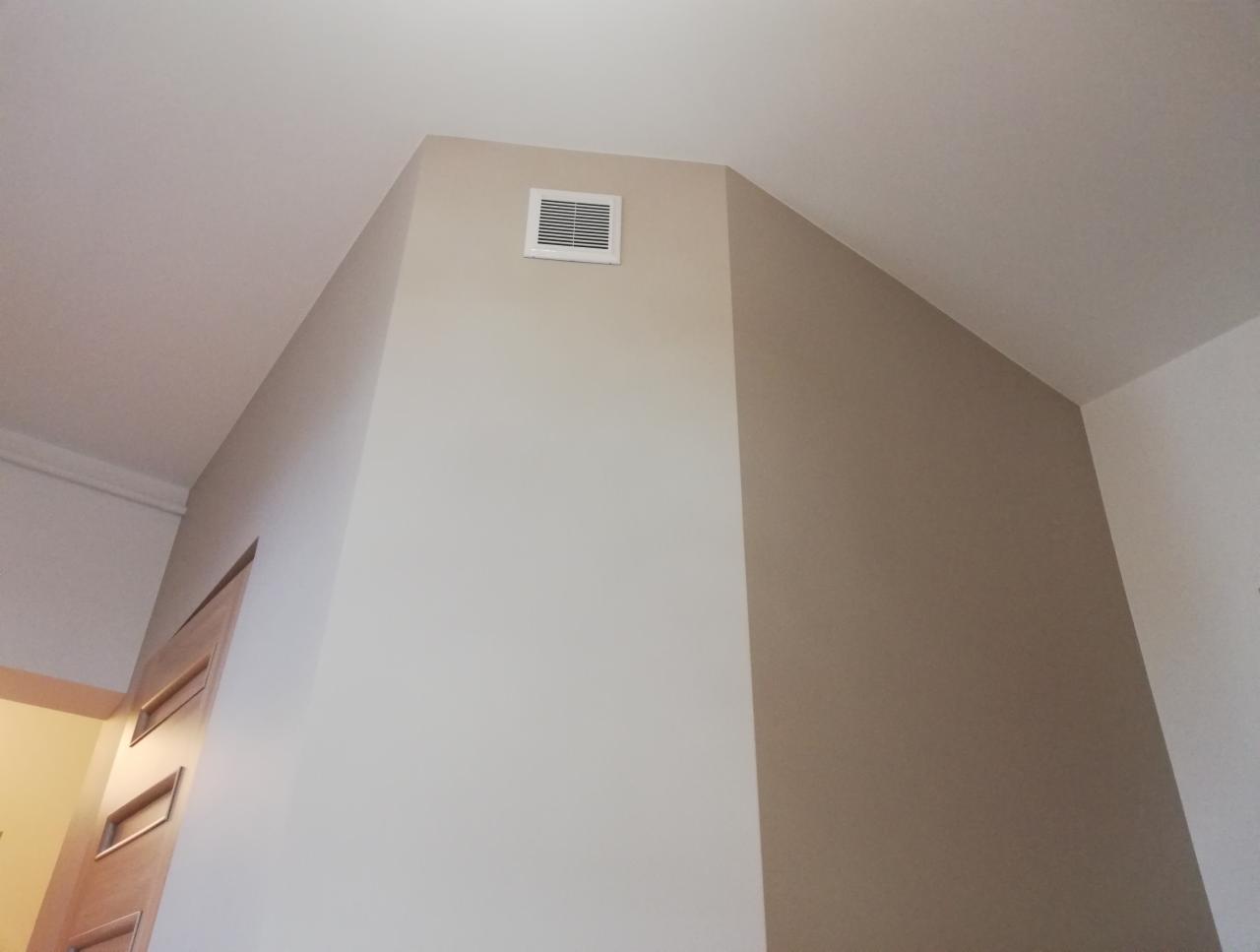 Wnętrze po remoncie: ściana pomalowana na dwa odcienie beżu, z kratką wentylacyjną pod sufitem i fragmentem drewnianych drzwi po lewej stronie.