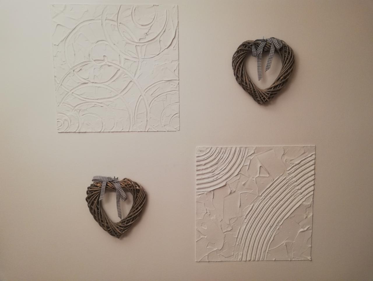 Dekoracyjna ściana z dwoma obrazami o fakturze gładzi szpachlowej i dwoma wiklinowymi sercami z szarą wstążką w kratę.