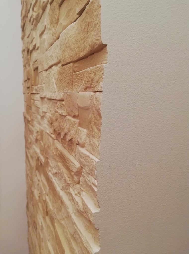 Fragment ściany z dekoracyjnym panelem imitującym kamień, sąsiadujący z gładką, pomalowaną ścianą w odcieniu beżu.