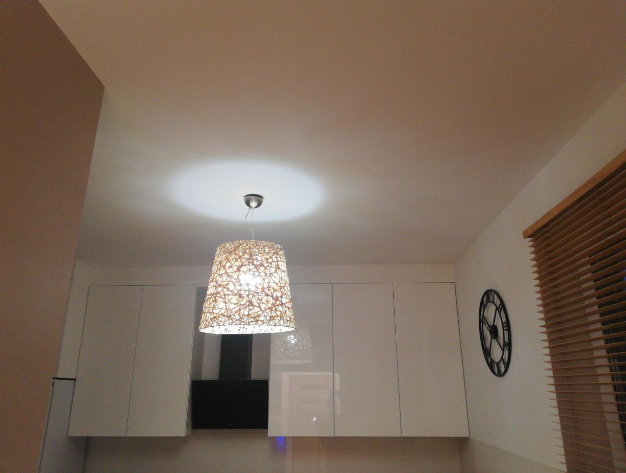 Wnętrze kuchni z białymi szafkami, designerską lampą wiszącą z abażurem z plecionki i zegarem ściennym w stylu retro, widoczne rolety drewniane w oknie.