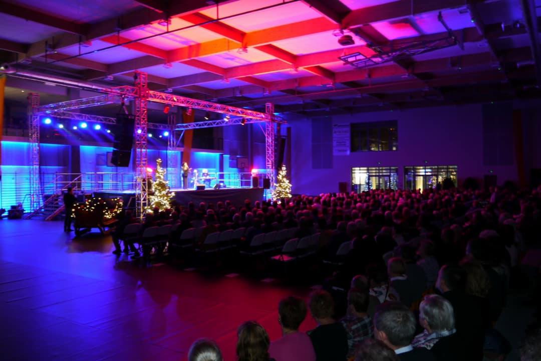 Widok na scenę z oświetleniem LED, dwie choinki i publiczność podczas wydarzenia w hali sportowej.