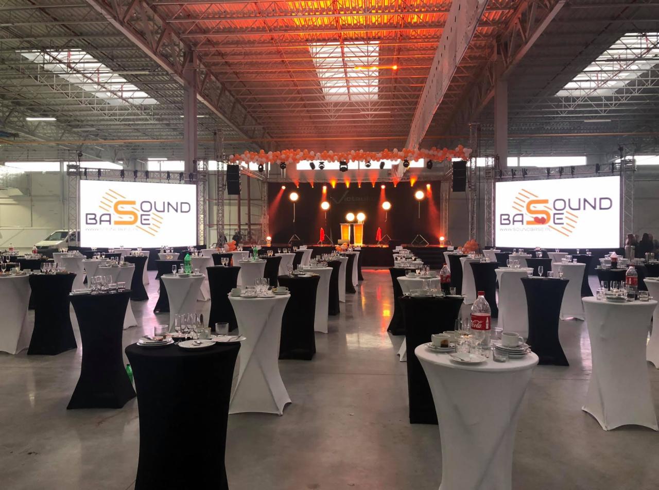 Sala bankietowa z ustawionymi stolikami koktajlowymi w czarno-białych pokrowcach, scena z oświetleniem i ekranami LED w tle, przygotowana na imprezę.