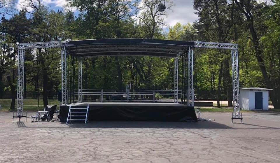 Scena plenerowa z metalową konstrukcją, zadaszeniem i schodami, ustawiona na betonowym placu w otoczeniu zieleni parkowej.