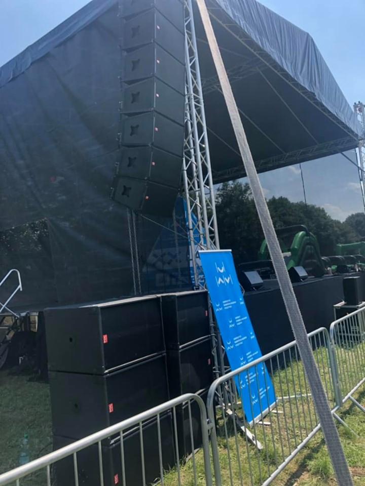 Scena plenerowa z nagłośnieniem line array, czarnym zadaszeniem i niebieskim banerem z logo za metalowym ogrodzeniem.