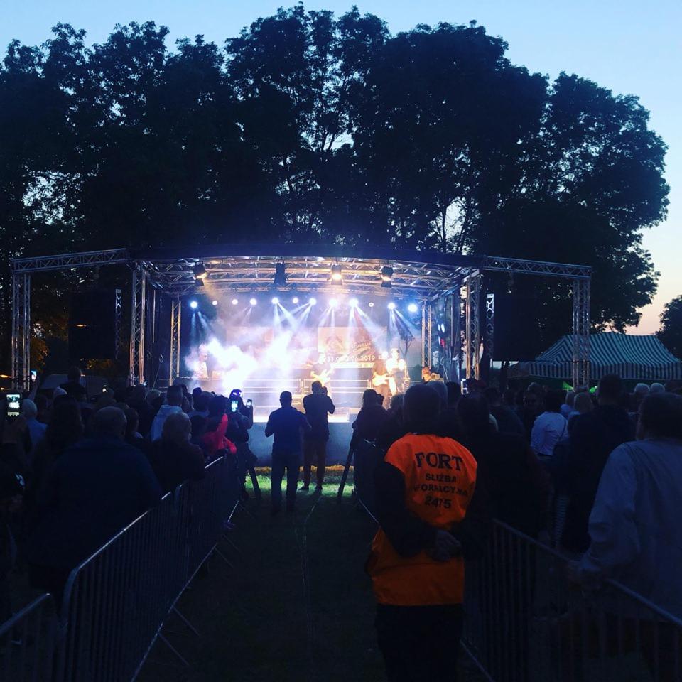 Wieczorny koncert plenerowy z oświetloną sceną, tłumem widzów i ochroną w pomarańczowej kamizelce z napisem 'FORT SŁUŻBA OCHRONY 24/5' na pierwszym planie.