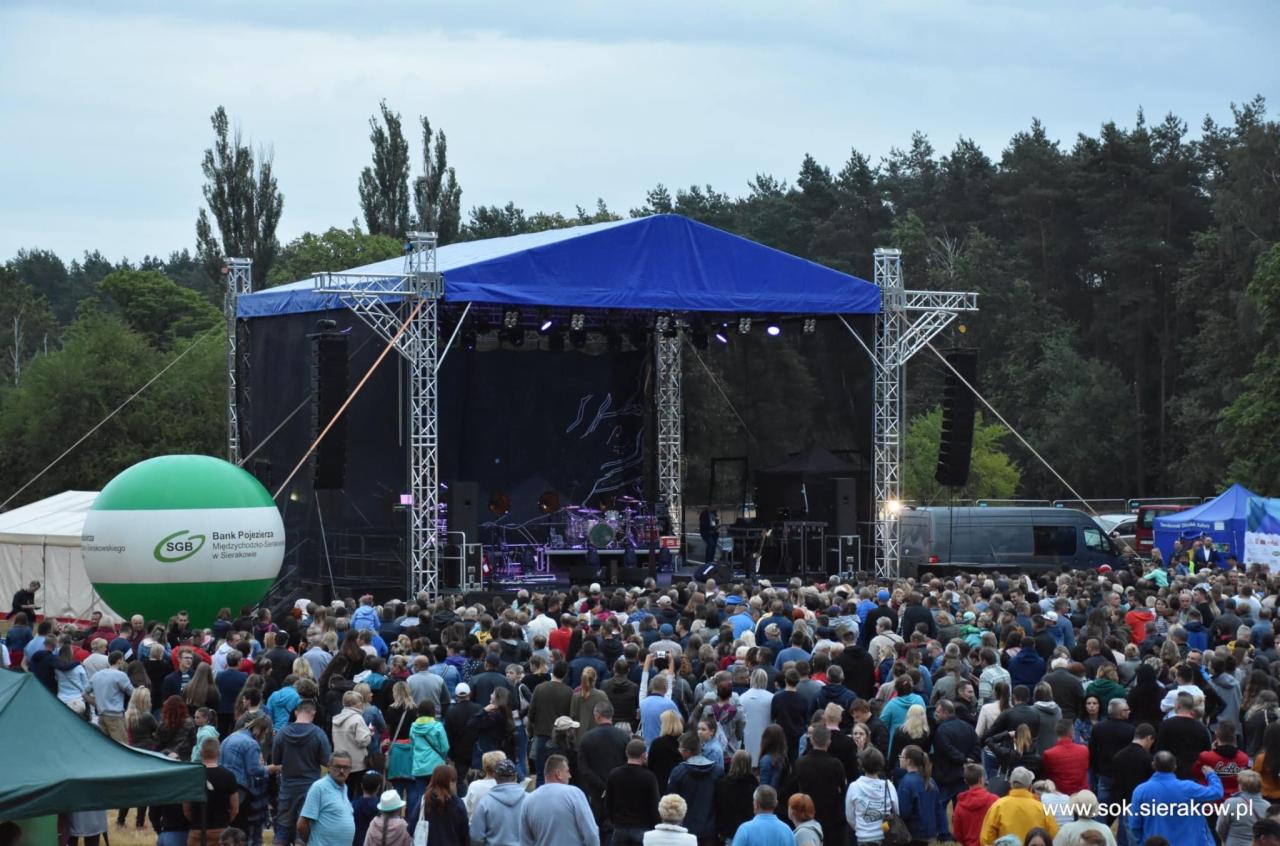 Plenerowy koncert z widoczną sceną z niebieskim zadaszeniem, tłumem ludzi i balonem reklamowym SGB w Sierakowie.