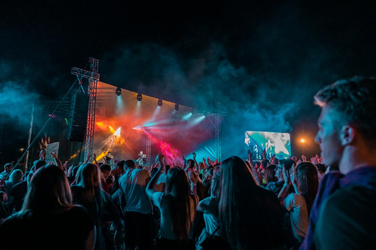 Tłum ludzi na koncercie z oświetloną sceną, efekty specjalne w postaci dymu i kolorowych świateł, widoczna konstrukcja sceny i ekran LED w tle.