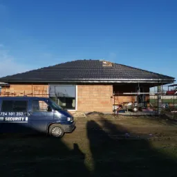 Dom w trakcie budowy z ciemną dachówką, częściowo ukończony dach, rusztowania i samochód dostawczy z napisem 'SECURITY' po lewej stronie.