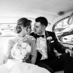 Czarno-białe zdjęcie pary młodej siedzącej w luksusowym samochodzie, z panną młodą trzymającą bukiet białych róż, oboje patrzą na siebie z czułością, widoczne krople deszczu na szybie.