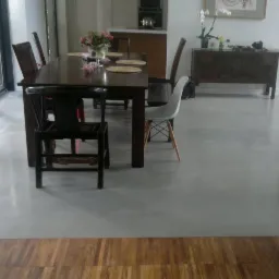 Jasnoszara, jednolita podłoga w nowoczesnym wnętrzu z dużym oknem, drewnianym stołem i krzesłami, przechodząca w drewnianą podłogę na pierwszym planie.