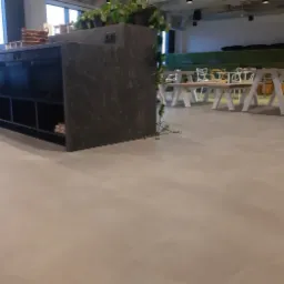 Jasnoszara, jednolita podłoga w przestronnym, nowoczesnym wnętrzu biurowym z roślinami i białymi stołami w tle.