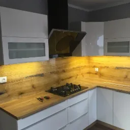 Nowoczesna kuchnia z białymi szafkami, drewnianym blatem i panelem ściennym z podświetleniem LED, widoczne narzędzia na blacie.