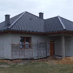 Dom jednorodzinny w stanie surowym zamkniętym z ciemną dachówką ceramiczną, widoczne rusztowania przy oknach i drzwiach, oraz stosy piasku i materiałów budowlanych na działce.