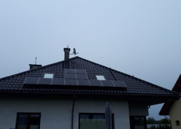 Panele słoneczne na ciemnym, spadzistym dachu domu jednorodzinnego z kominem i anteną satelitarną, pochmurne niebo w tle.