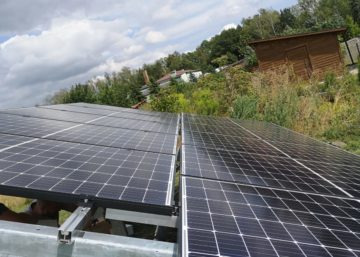 Panele słoneczne na dachu z widocznym systemem montażowym, w tle zieleń i zabudowania, pochmurne niebo.