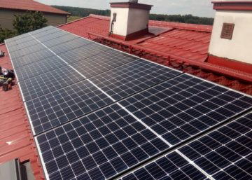 Panel słoneczny na czerwonym dachu z kominem w tle, widok z góry, częściowo widoczne narzędzia monterskie po lewej stronie.