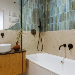 Nowoczesna łazienka z wanną obudowaną płytkami, umywalką nablatową na drewnianej szafce, okrągłym lustrem i armaturą w kolorze czarnym.