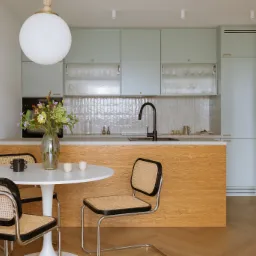 Jasna kuchnia z pastelowymi szafkami, drewnianą wyspą, okrągłym białym stołem z rattanowymi krzesłami i bukietem kwiatów w szklanym wazonie.