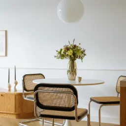 Jasne wnętrze jadalni z okrągłym, białym stołem, bukietem w szklanym wazonie, trzema krzesłami z rattanowymi siedziskami i oparciami oraz lampą kulą zwisającą z sufitu. Na komodzie widoczne wysokie...