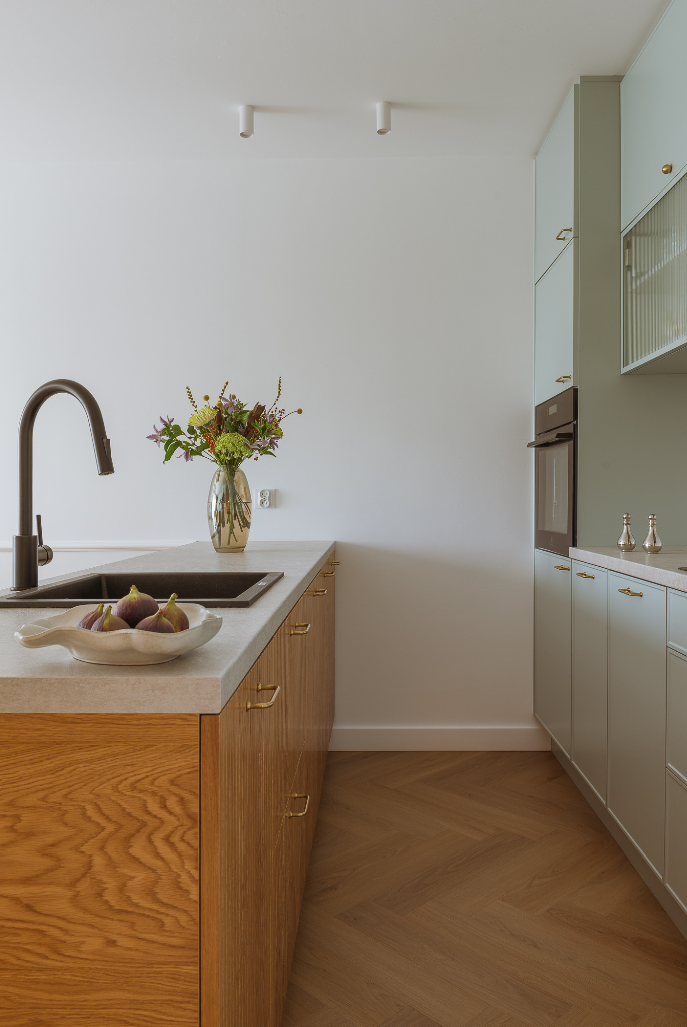 Nowoczesna kuchnia z drewnianymi frontami szafek i blatem z konglomeratu, czarny zlew, misa z figami, wazon z kwiatami, podłoga z parkietu ułożonego w jodełkę.