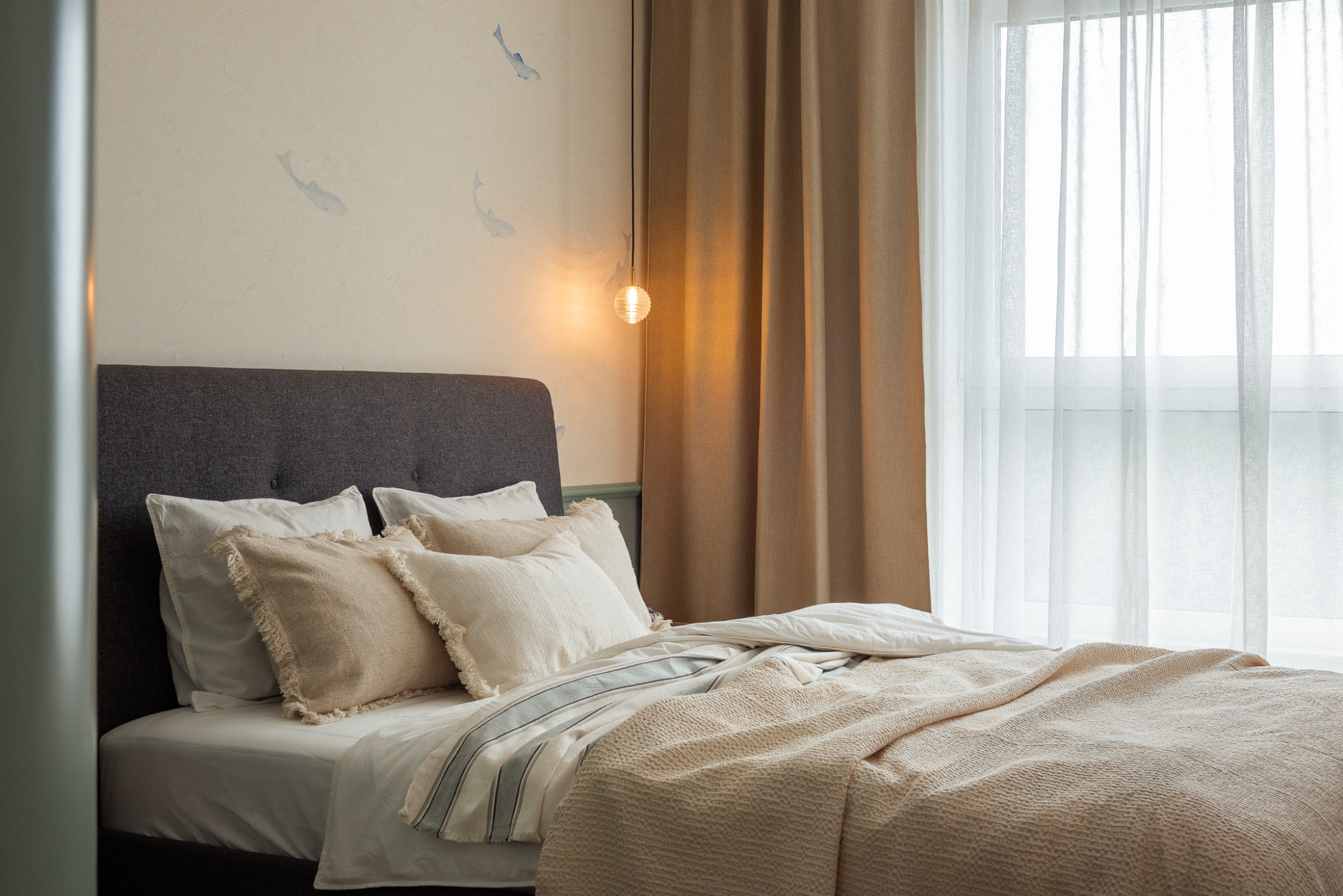 Jasna sypialnia z łóżkiem z szarym wezgłowiem, beżowymi poduszkami z frędzlami, narzutą w wafelkowy wzór oraz lampą wiszącą z kulistym kloszem, na ścianie subtelny motyw ryb